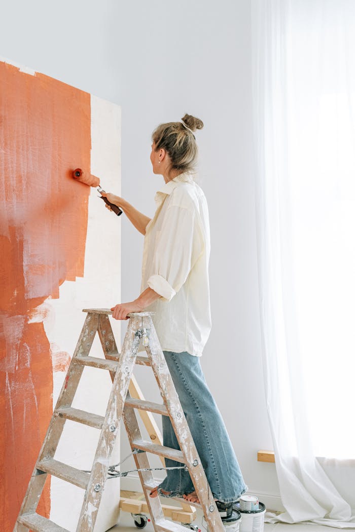 An adult woman painting a wall with red paint using a roller indoors, standing on a ladder.