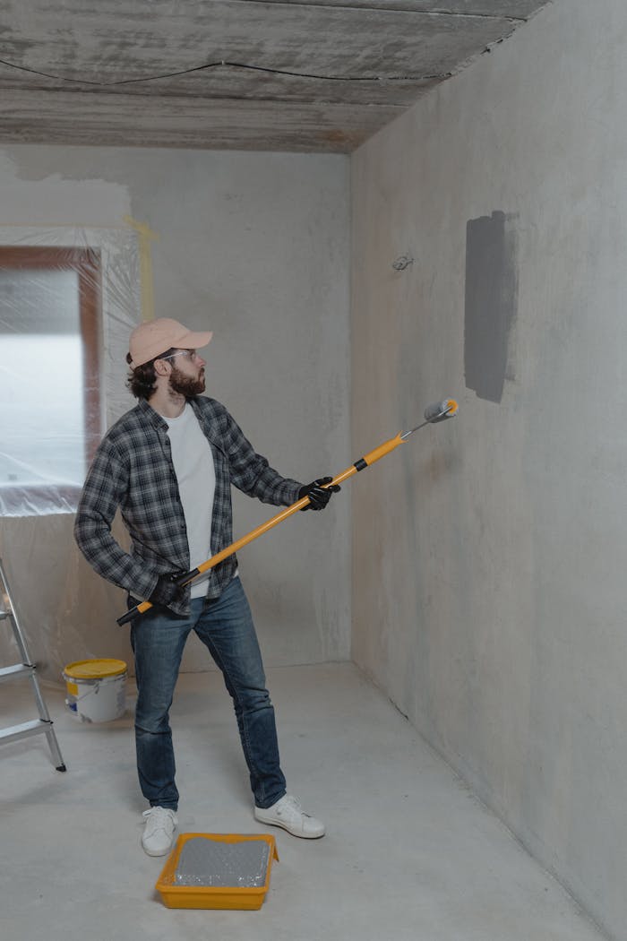 A man paints a wall gray in a room under renovation, using a roller for a smooth finish.